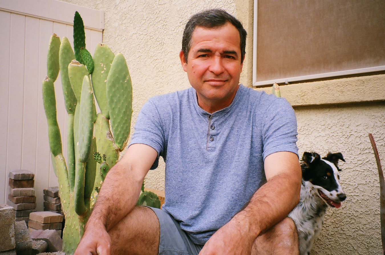 Emmanuel's father sitting, facing forward and looking at the camera happily. A black and white dog sits next to him calmly with a green cactus in the background.